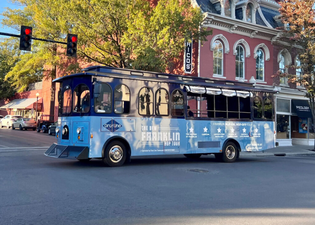 All Aboard the Franklin Hop! How to Experience This Unique Sightseeing Trolley Tour A blue trolley bus with 'Franklin Hop-On Hop-Off Tour' signage travels down a city street in Williamson County, Tennessee.