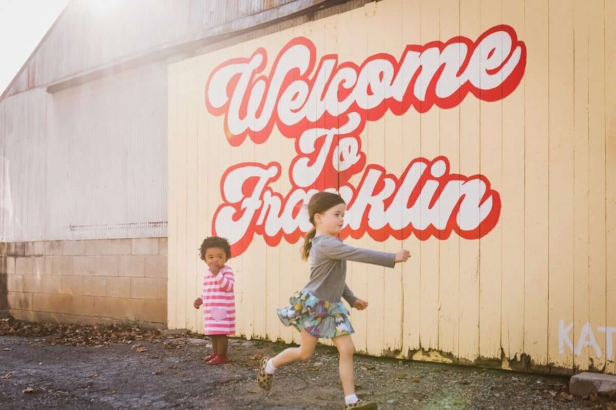 Two young girls playfully run past a large, colorful 'Welcome to Franklin' mural in Williamson County, Tennessee.
