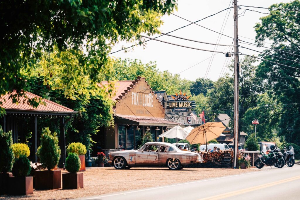 A vintage car is parked outside a live music venue with outdoor seating and umbrellas in Williamson County, Tennessee.
