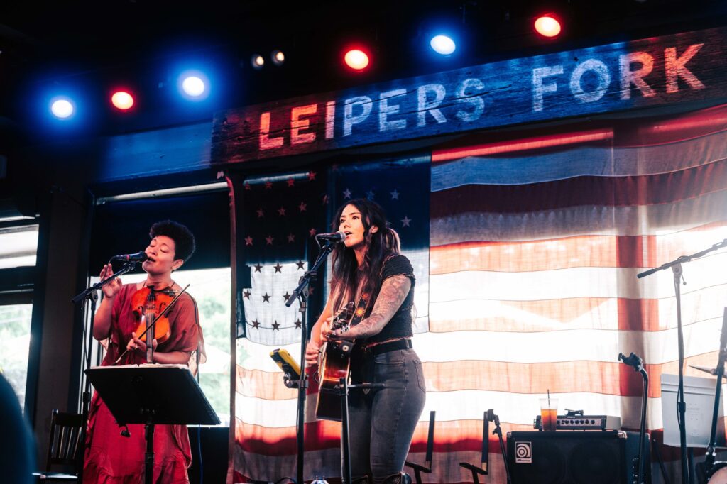 Two women perform on stage with microphones and instruments, backed by an American flag in Williamson County, Tennessee.