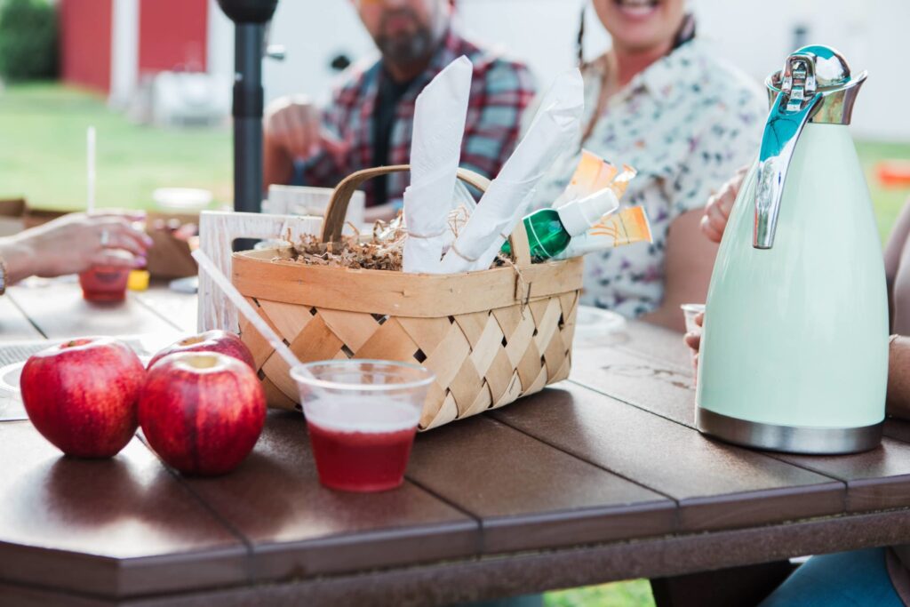 A picnic table with a basket of apples, a pitcher of red drink, and a green thermos in Williamson County, Tennessee.