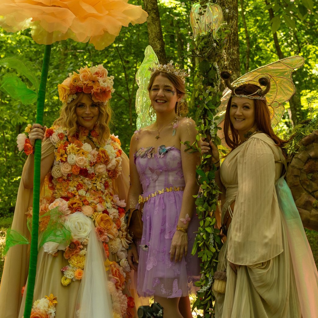 Three women dressed as fairies, adorned with vibrant floral crowns and wings, stand together in a lush forest, smiling and holding a large orange flower and a staff with glowing orbs in Williamson County, Tennessee.