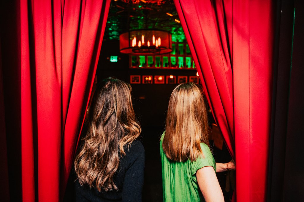 Two women with long hair stand behind red curtains, looking into a dimly lit room with a chandelier and green lighting in Williamson County, Tennessee.