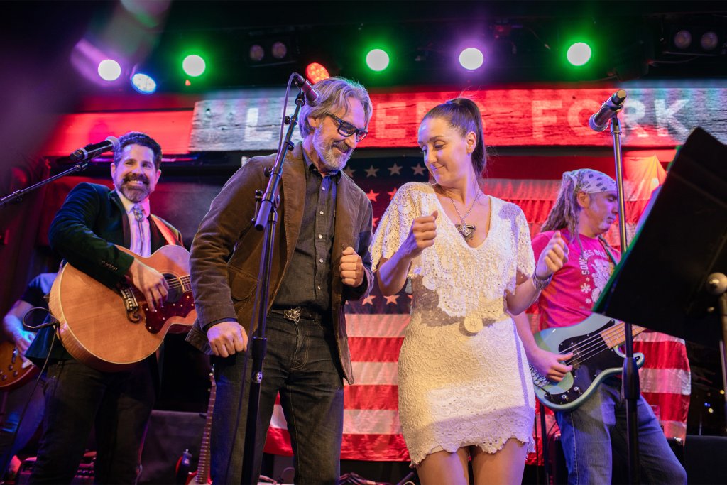A lively band performs on stage, featuring a man in a brown jacket playing an acoustic guitar, a woman in a white dress singing into a microphone, and two other musicians with electric guitars, all set against a backdrop of American flags and colorful stage lights in Williamson County, Tennessee.