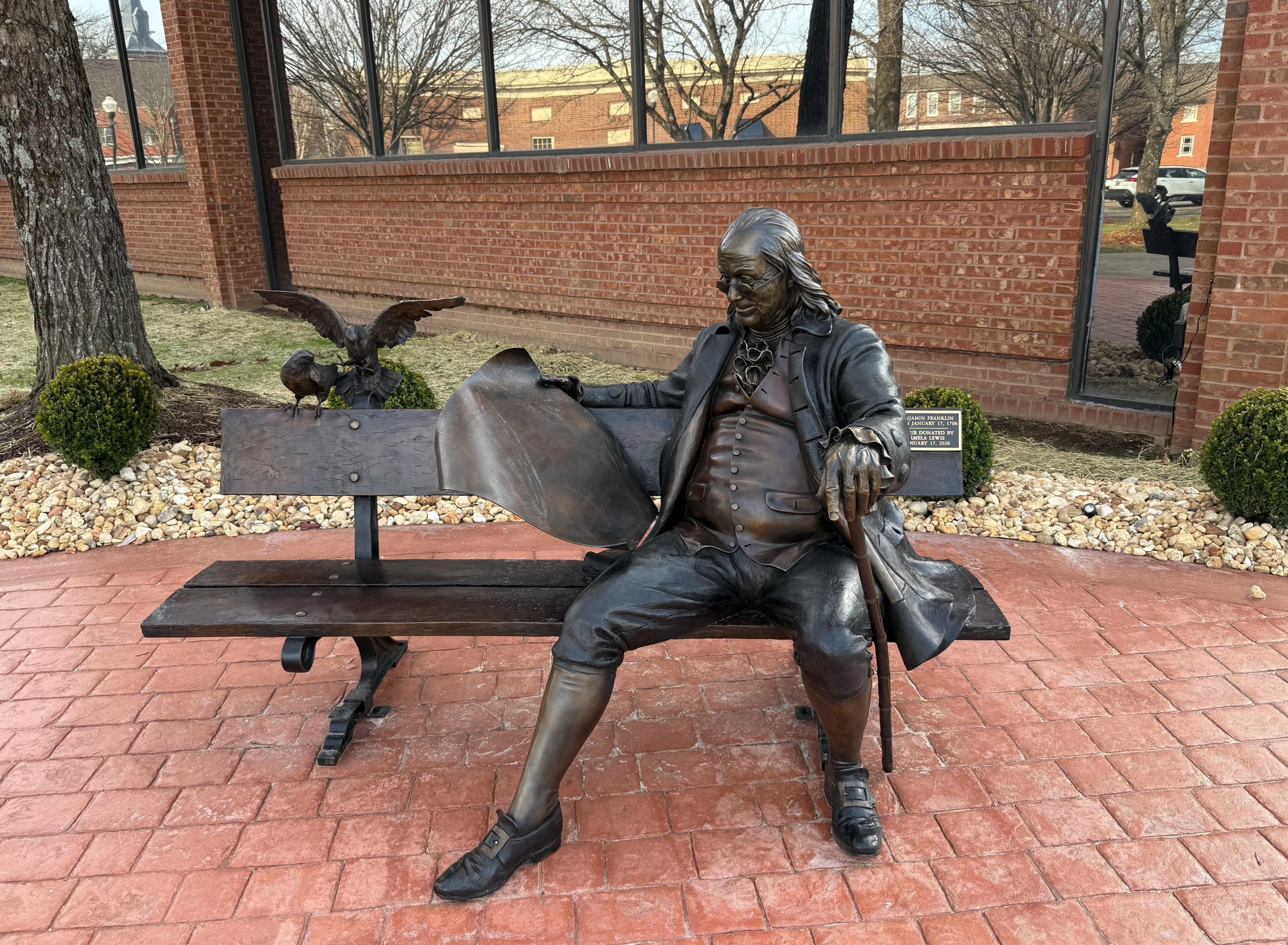 A bronze statue of Benjamin Franklin seated on a bench, holding a book and accompanied by an eagle and a turtle, set against a backdrop of a brick building and landscaped garden in Williamson County, Tennessee.