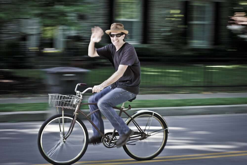 A man wearing a hat and sunglasses waves while riding a bicycle with a basket in Williamson County, Tennessee.