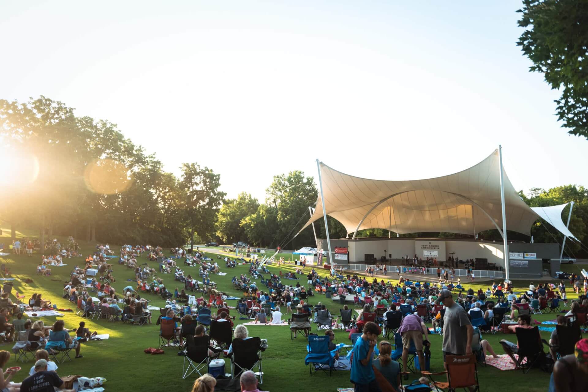 A large crowd enjoys an outdoor concert under a white tent at sunset in a grassy park in Williamson County, Tennessee.