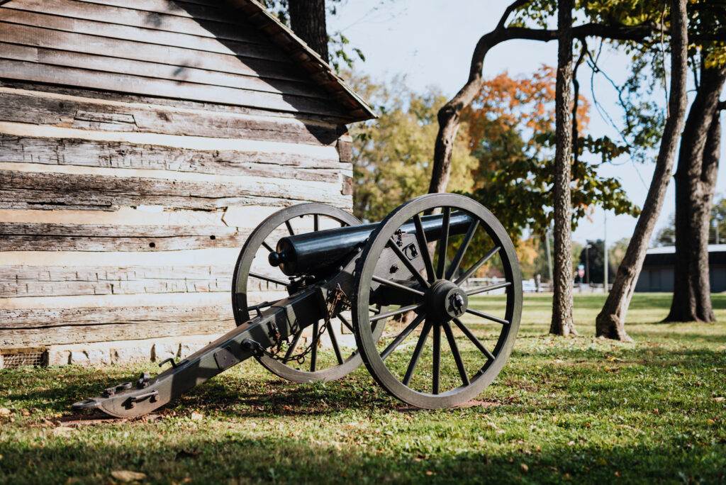 An old cannon with large wheels rests on grass beside a rustic wooden cabin in Williamson County, Tennessee.