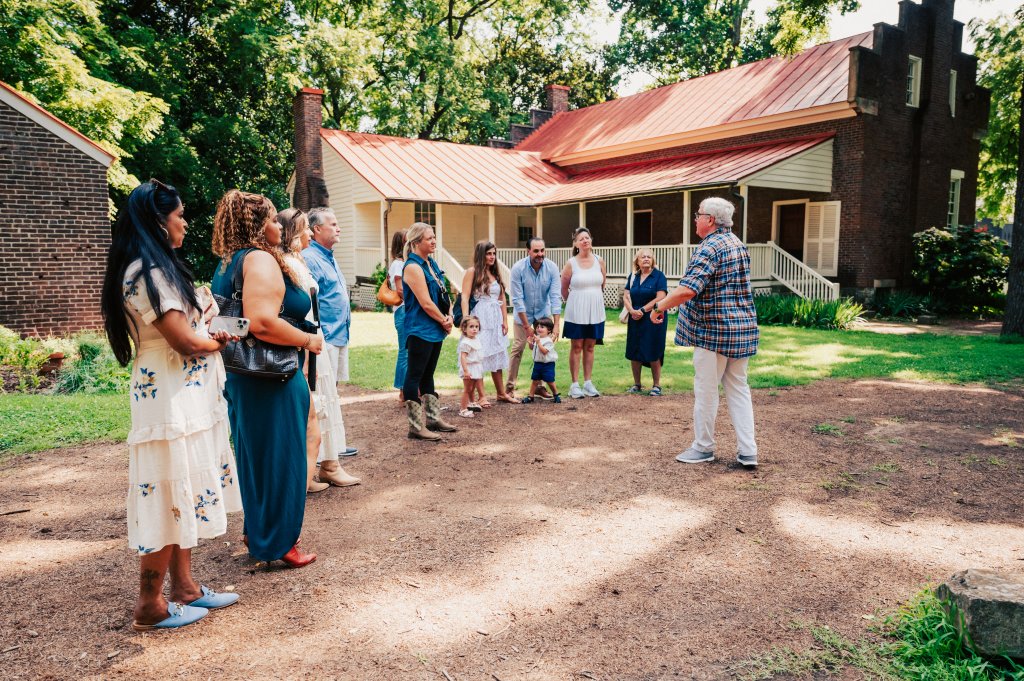 A diverse group of people, including adults and children, gather in a circle on a dirt path in front of a historic brick house with a red roof, surrounded by lush greenery and gardens in Williamson County, Tennessee.