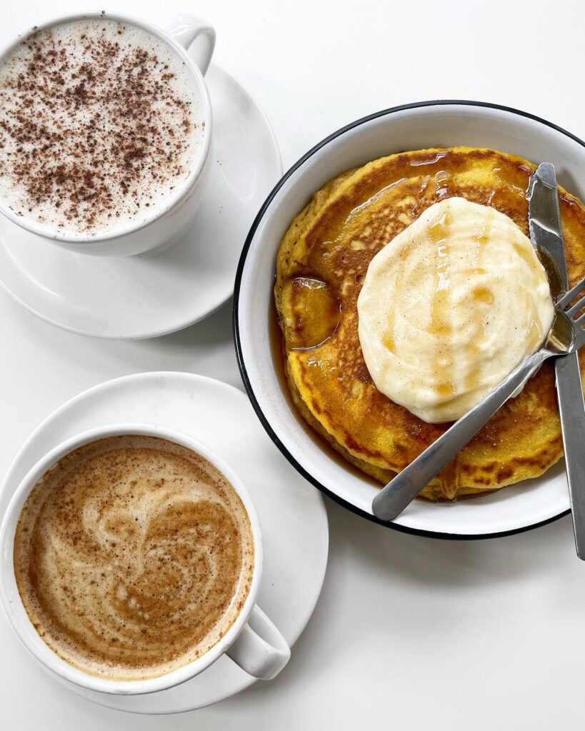 A bowl of golden pancakes topped with whipped cream and syrup, accompanied by two cups of coffee with foam art in Williamson County, Tennessee.