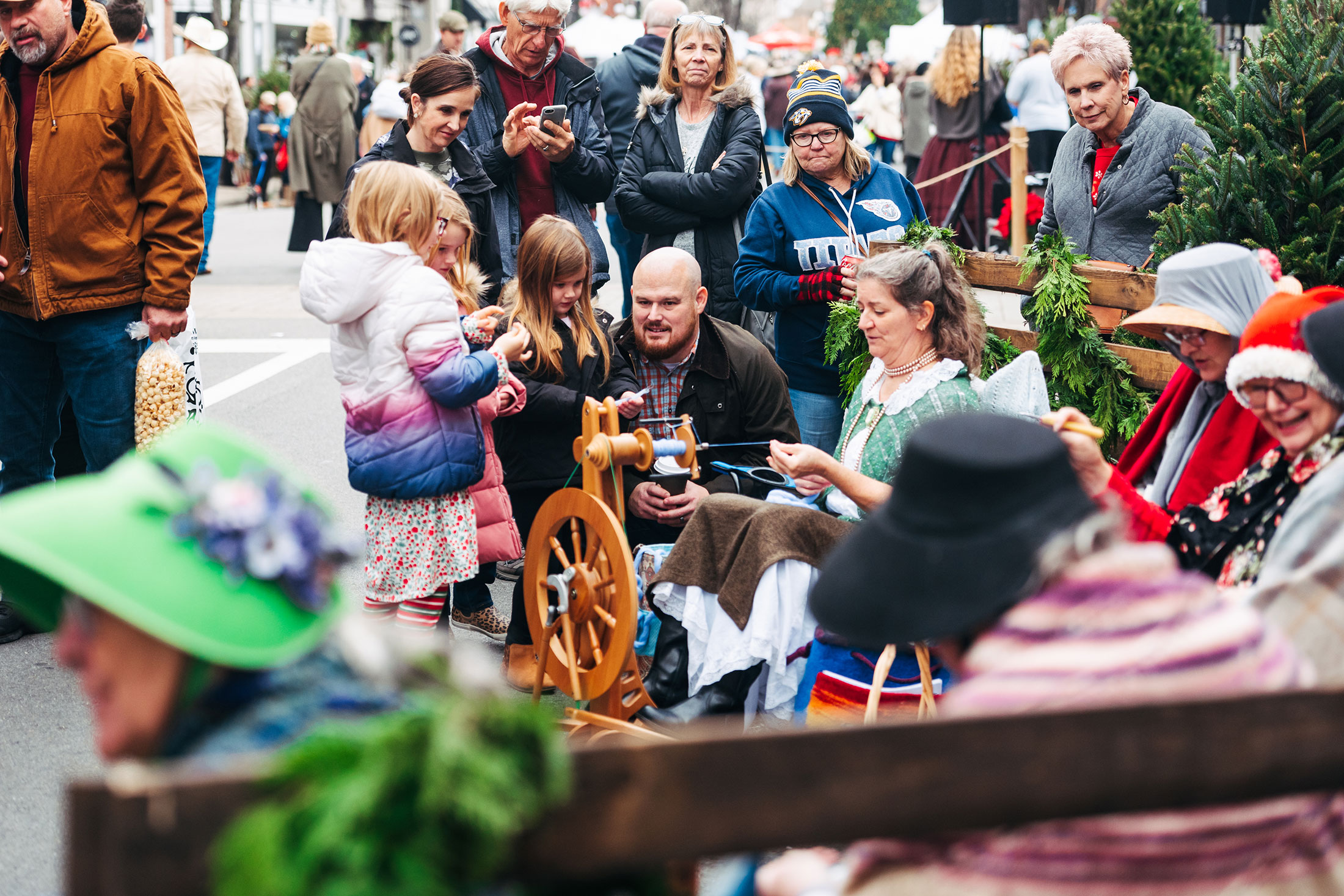A lively holiday market scene with people gathered around a spinning wheel, children interacting with adults, and festive decorations including greenery and a Christmas tree in Williamson County, Tennessee.