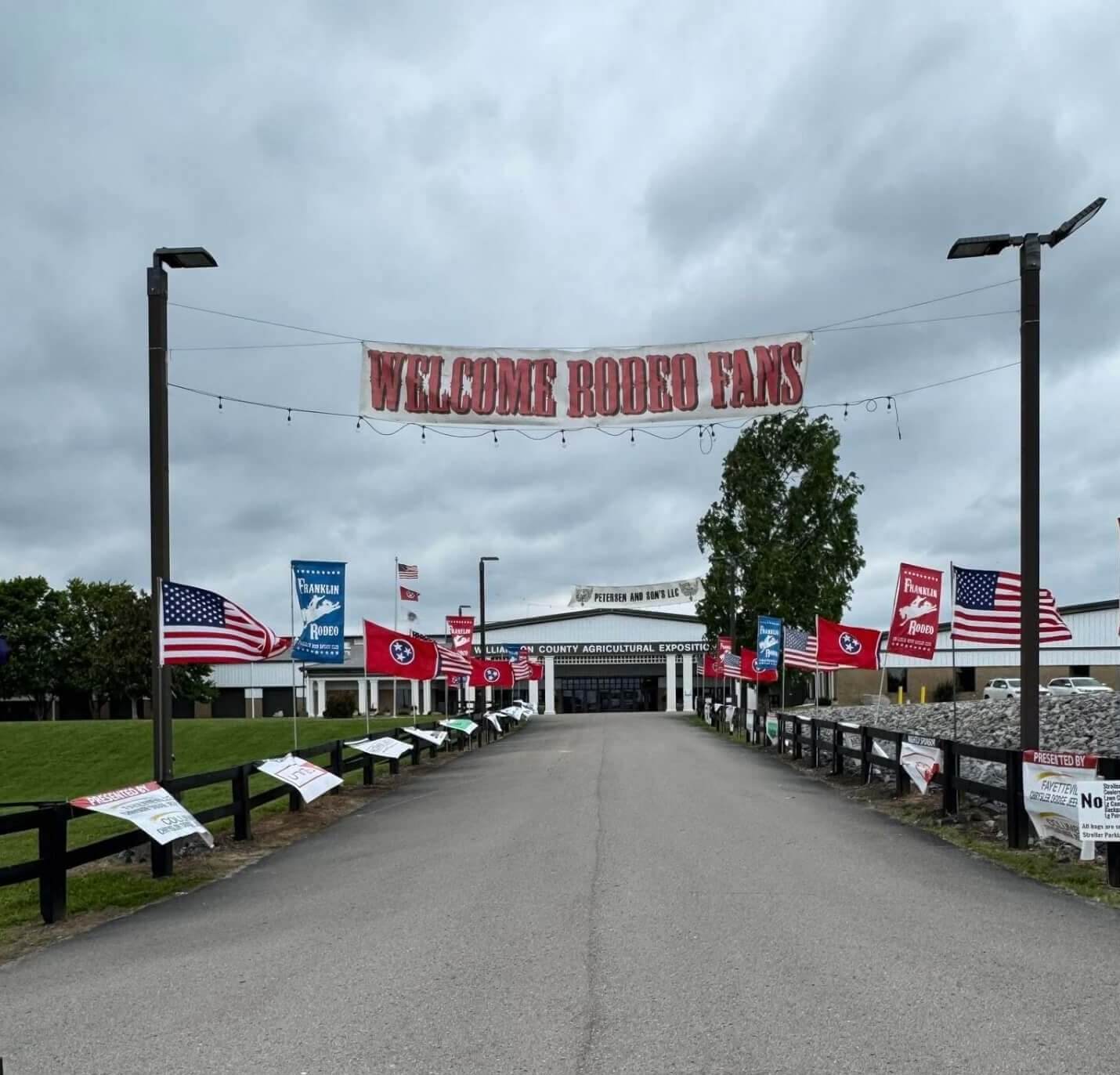 The Franklin Rodeo A wide, empty road leads towards a large building adorned with numerous flags, including American and rodeo-themed banners, under a cloudy sky in Williamson County, Tennessee.