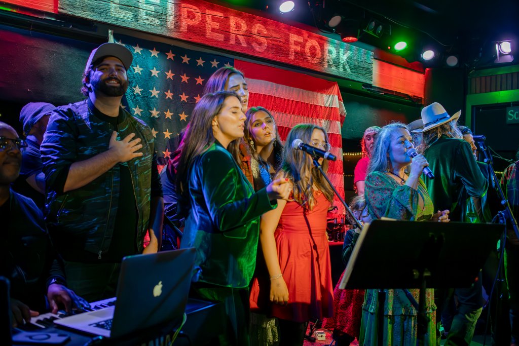 A lively band performs on stage, with a diverse group of musicians singing and playing instruments under a backdrop of an American flag and neon signs in Williamson County, Tennessee.