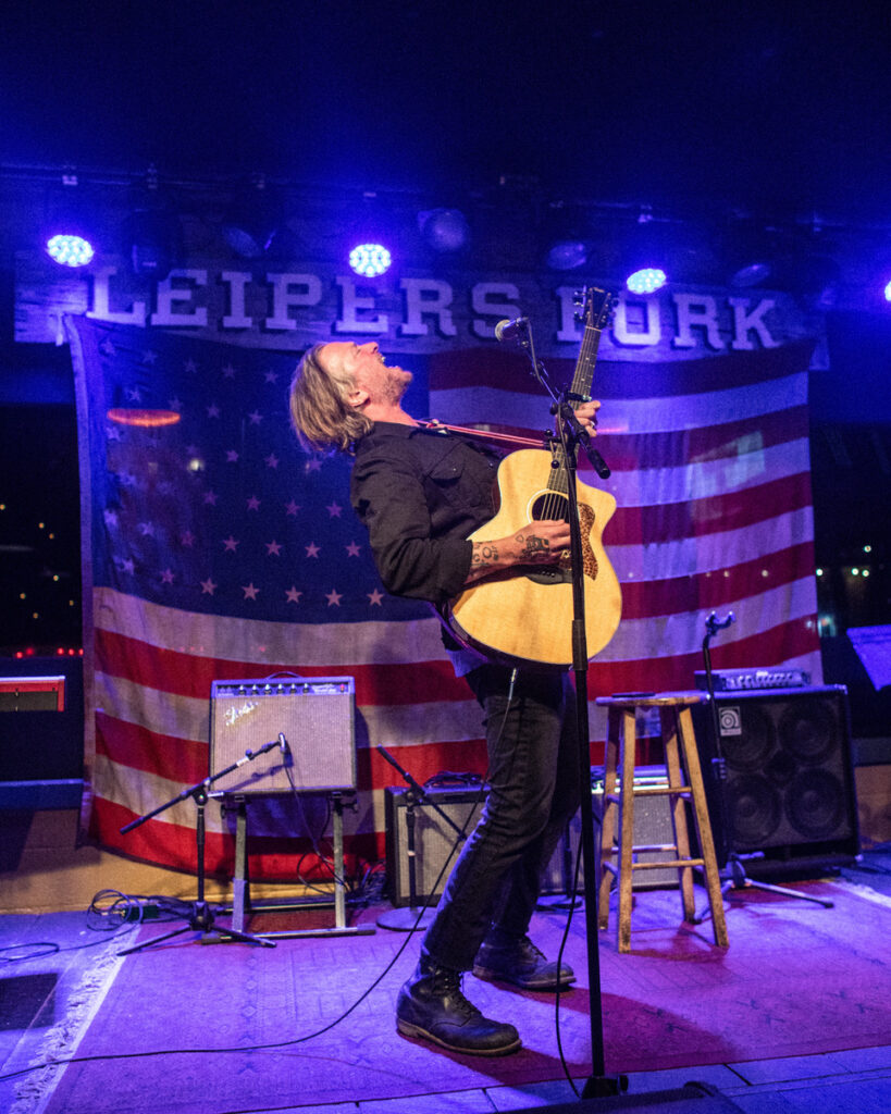 A musician passionately plays an acoustic guitar on stage, backed by an American flag in Williamson County, Tennessee.