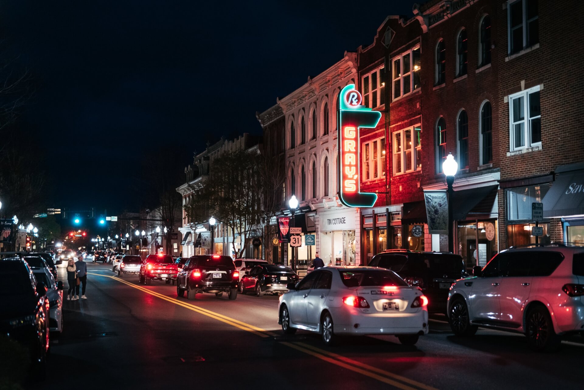 A bustling downtown street at night, illuminated by neon signs and streetlights, with cars driving and pedestrians walking in Williamson County, Tennessee.