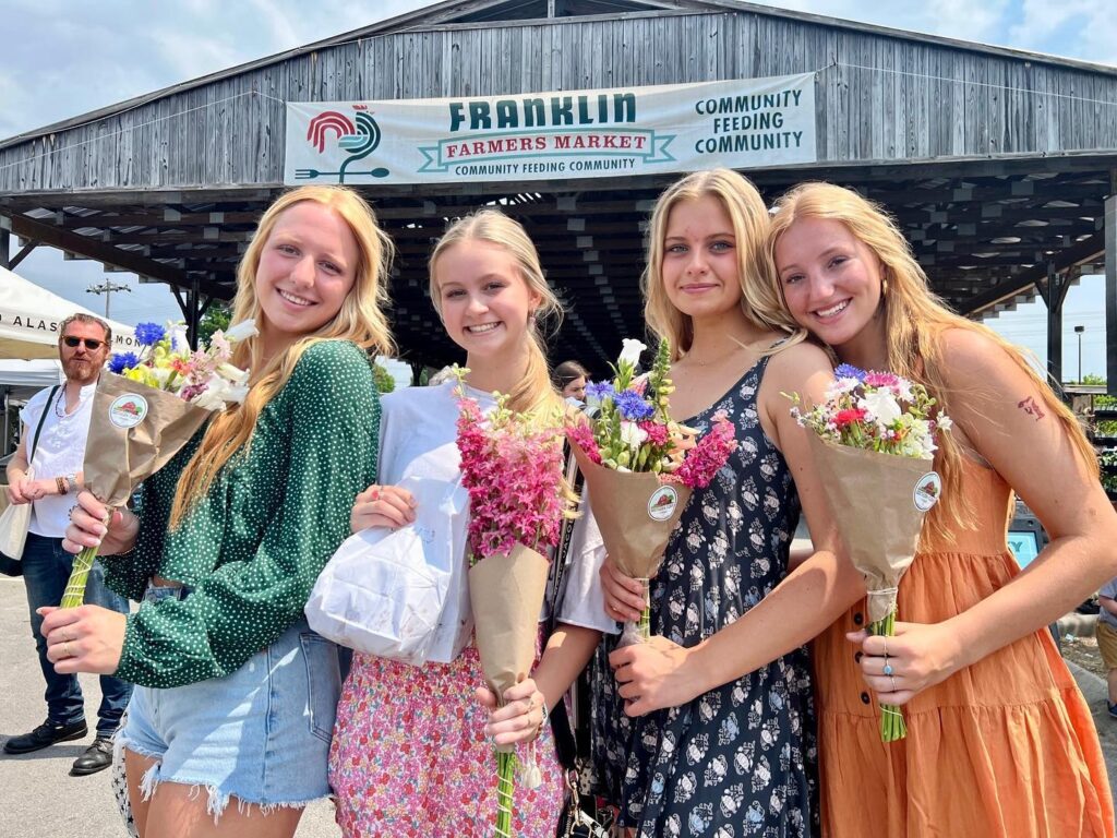 Four young women smiling and holding bouquets of flowers at a farmers market in Williamson County, Tennessee.