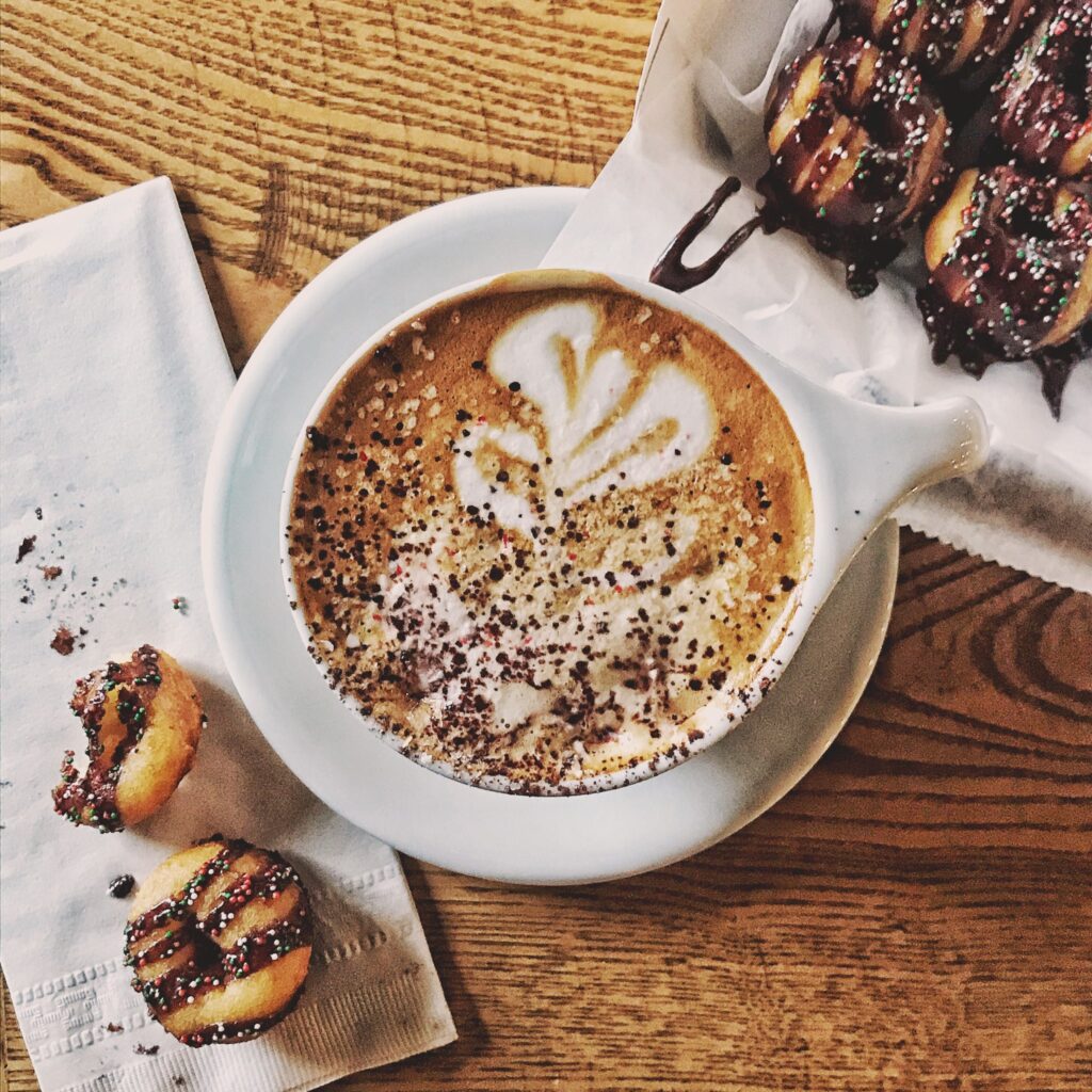 A steaming cup of coffee with latte art sits on a wooden table next to chocolate-dipped doughnuts in Williamson County, Tennessee.