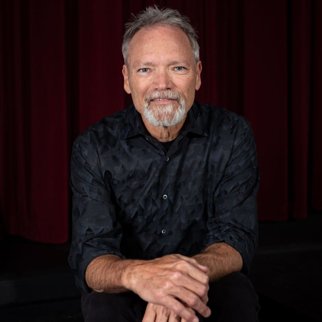 A man with gray hair and a beard, wearing a dark patterned shirt, sits with his hands clasped in front of him against a red curtain backdrop in Williamson County, Tennessee.
