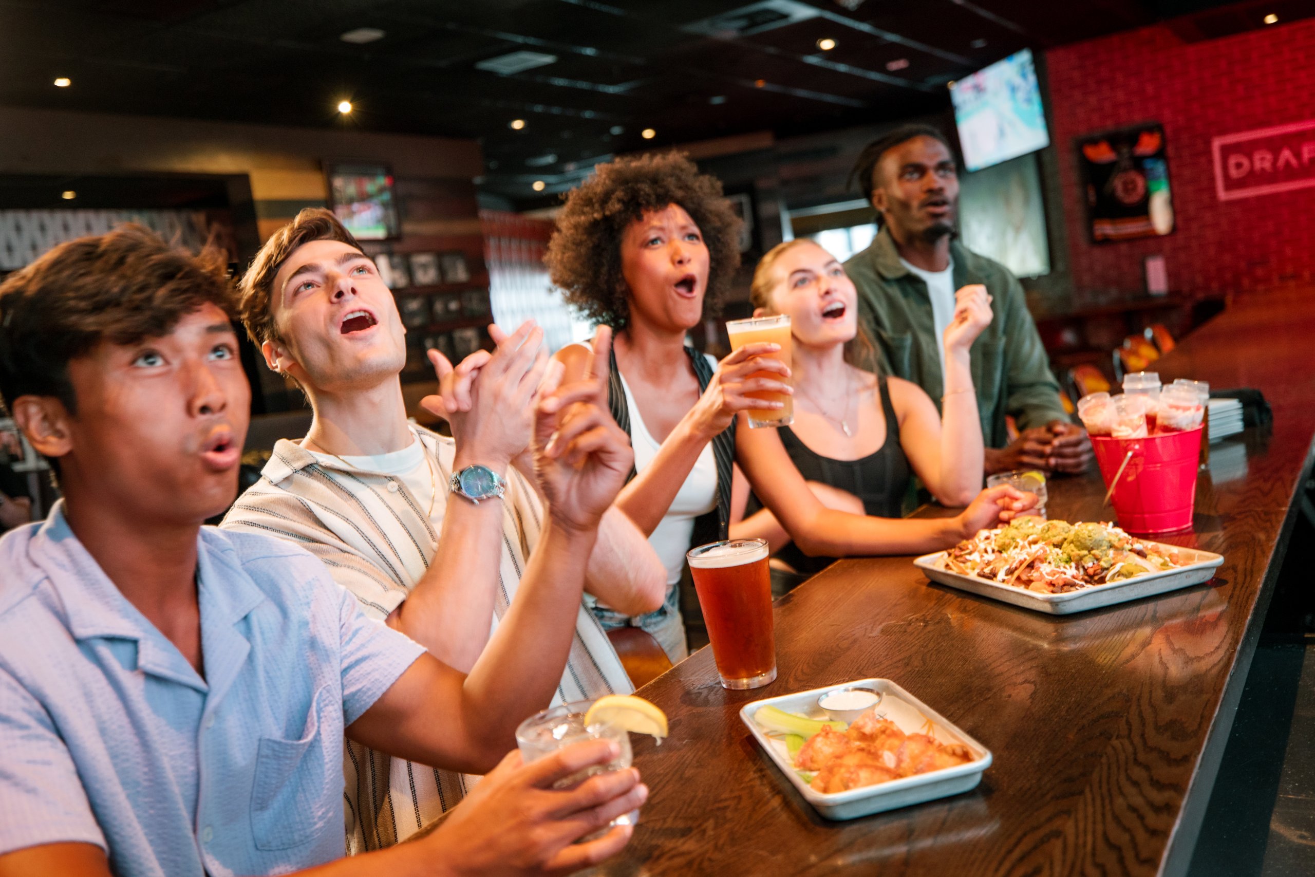 A group of five friends, three men and two women, are gathered around a bar table, cheering and clapping while watching a sports game on a TV screen above them. They are enjoying various drinks and snacks, including beer, margaritas, and plates of nachos and chicken wings in Williamson County, Tennessee.