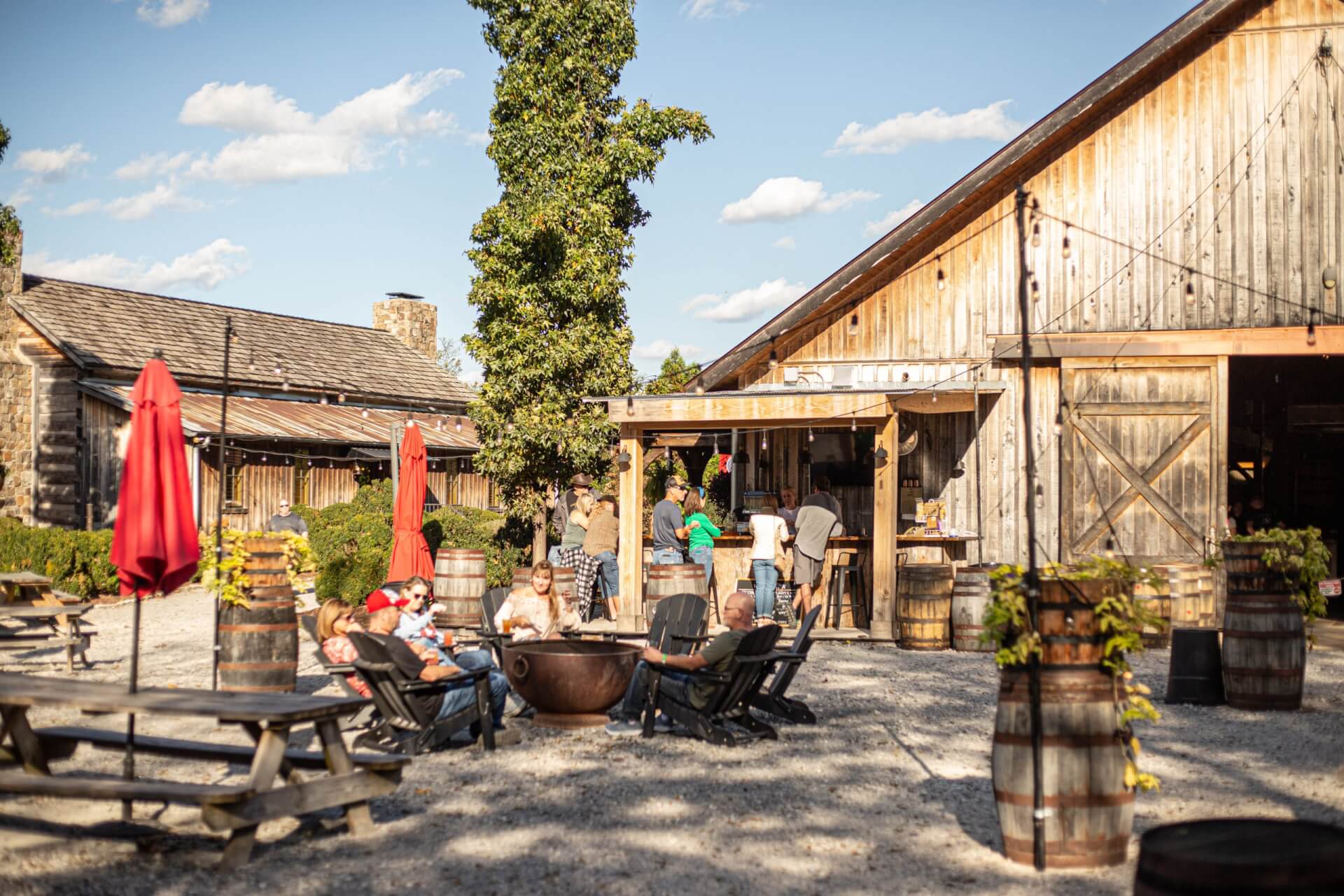 A rustic outdoor gathering area with wooden picnic tables, Adirondack chairs, and a fire pit surrounded by people enjoying the sunny day in Williamson County, Tennessee.