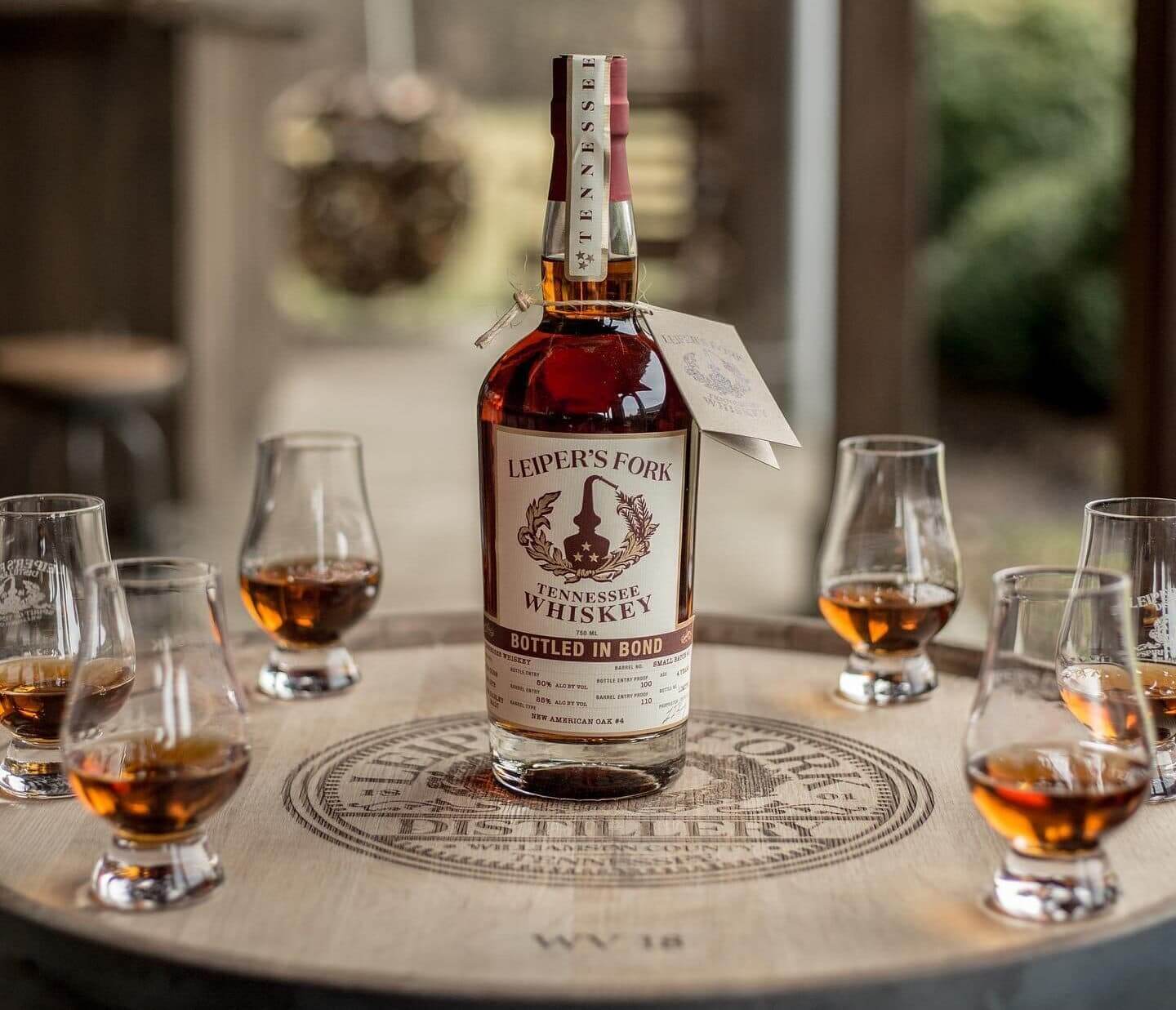 A bottle of Leiper's Fork Tennessee whiskey sits on a wooden barrel surrounded by small glasses filled with amber liquid in Williamson County, Tennessee.