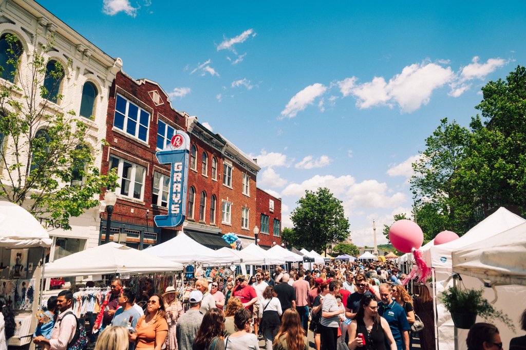 A bustling outdoor market fills a town square with white tents, shoppers, and colorful balloons against a backdrop of historic brick buildings and a clear blue sky in Williamson County, Tennessee.