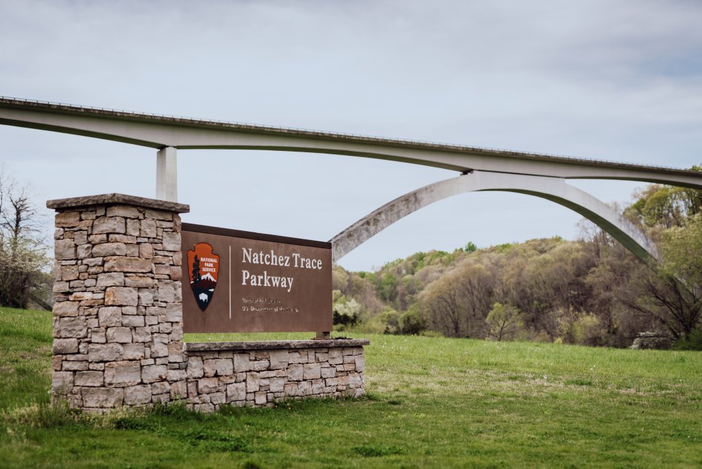 A stone sign for Natchez Trace Parkway stands beneath an arched bridge, surrounded by lush green grass and trees in Williamson County, Tennessee.