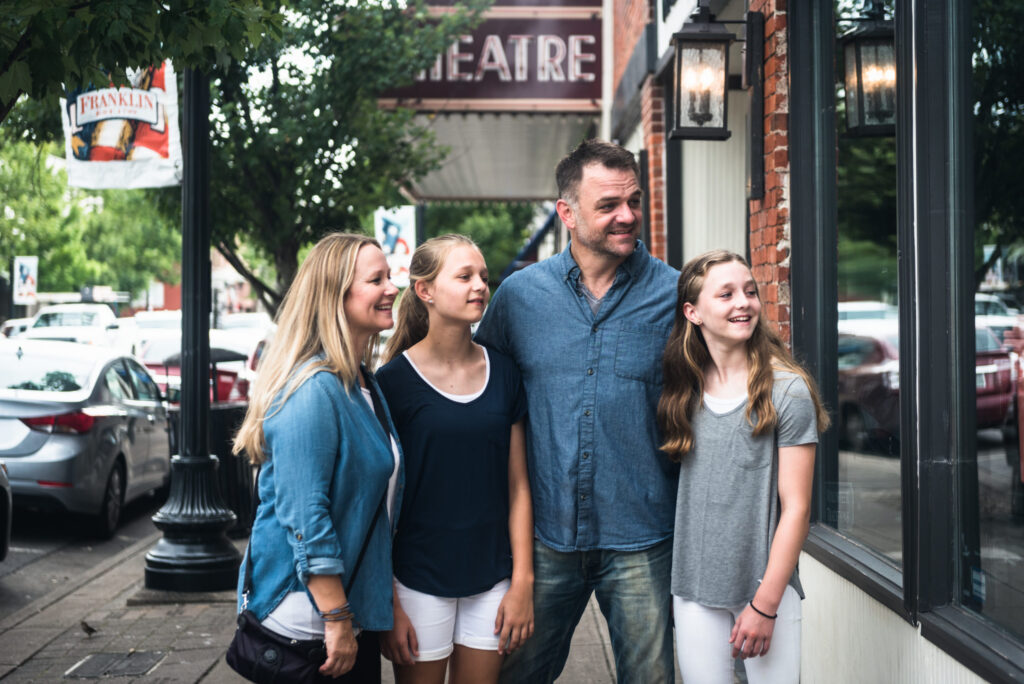 A family of four, including two girls and a boy, are walking down a street lined with shops and cars in Williamson County, Tennessee.