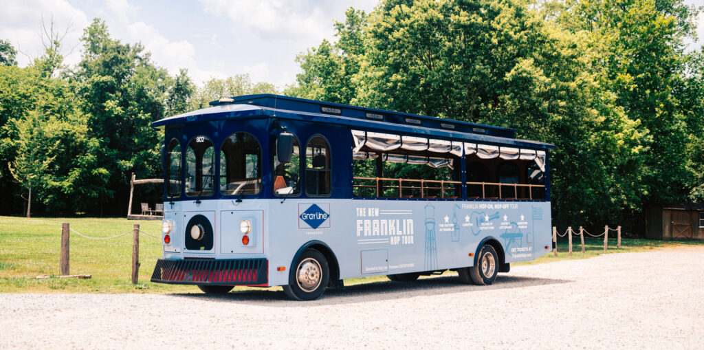 A blue trolley bus with 'Franklin Hop Tour' branding is parked on a gravel road surrounded by trees in Williamson County, Tennessee.
