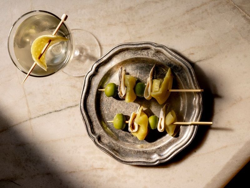 A martini glass with a lemon twist and a plate of stuffed olives and pickles on a marble surface in Williamson County, Tennessee.