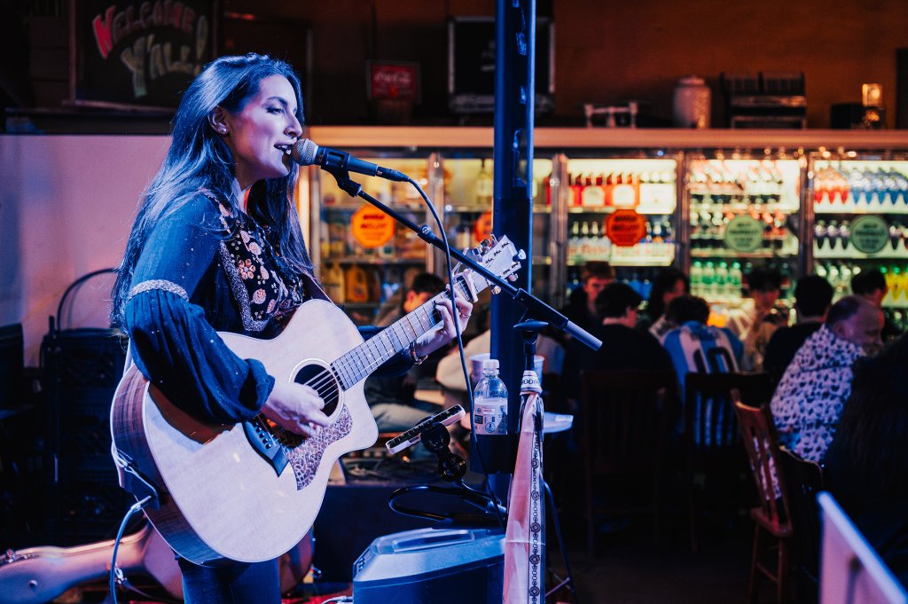 A woman with long dark hair performs an acoustic set in a dimly lit bar, singing into a microphone while playing an acoustic guitar in Williamson County, Tennessee.