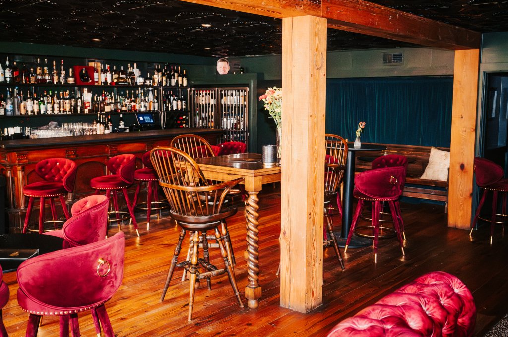 A dimly lit, cozy bar with a rustic wooden interior, featuring a variety of plush red velvet chairs and stools, a central wooden table, and a well-stocked bar area with shelves of liquor bottles in Williamson County, Tennessee.