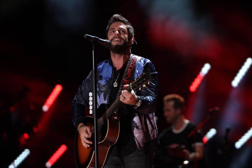 A musician performs on stage, strumming an acoustic guitar and singing into a microphone in Williamson County, Tennessee.