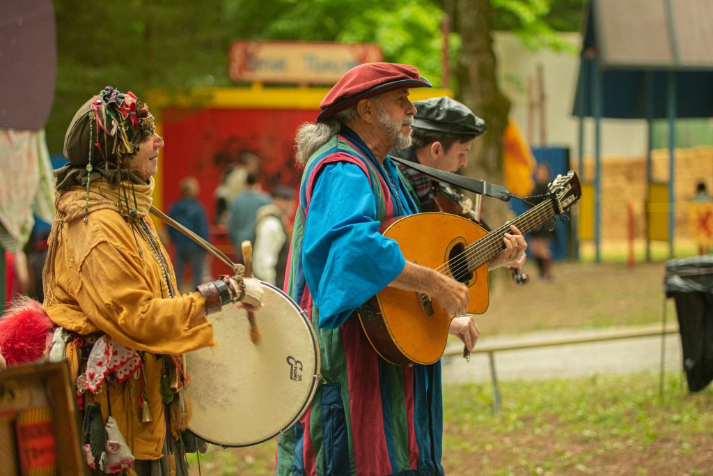 Two musicians dressed in colorful, traditional costumes play a drum and guitar in an outdoor setting, surrounded by a crowd and a festive atmosphere in Williamson County, Tennessee.