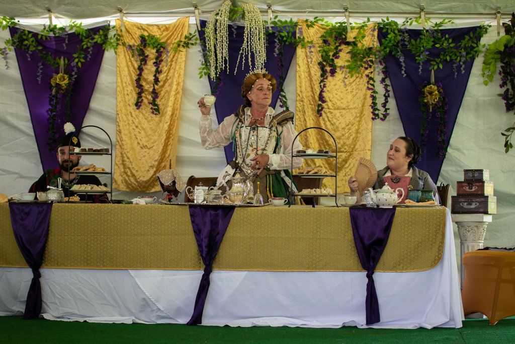 A woman dressed in a regal costume stands at a lavishly decorated table, holding a teacup, with two other costumed individuals seated beside her in Williamson County, Tennessee.
