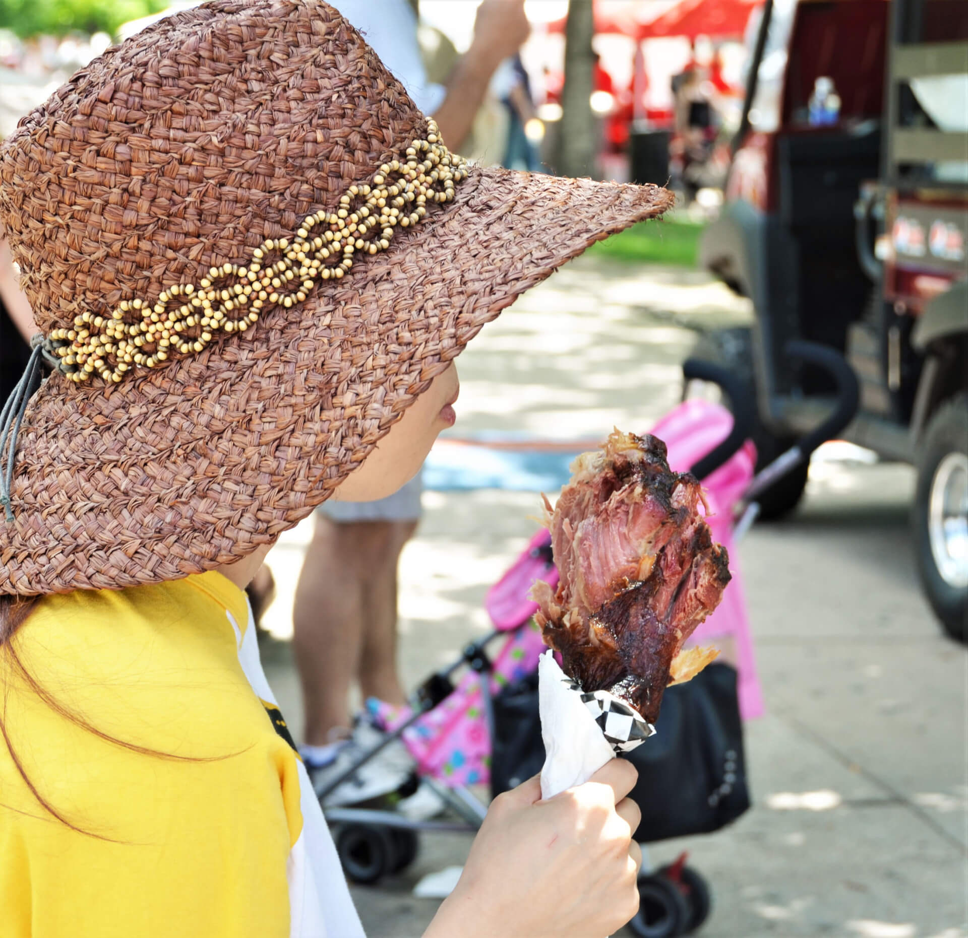 A young girl wearing a wide-brimmed straw hat with a beaded band enjoys a large, juicy drumstick at an outdoor event in Williamson County, Tennessee.