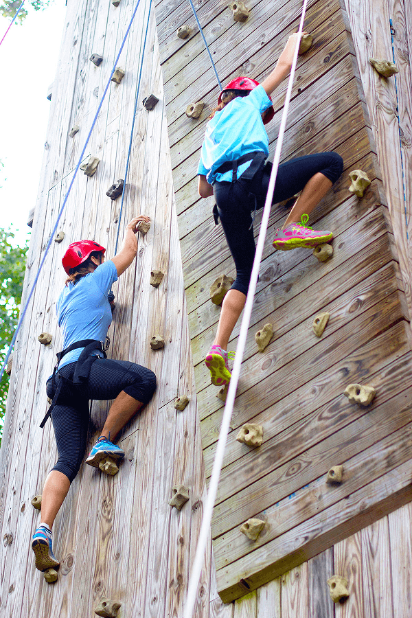 Two women in helmets and athletic wear climb a wooden rock climbing wall outdoors in Williamson County, Tennessee.