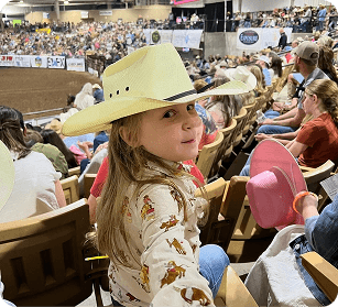 The Franklin Rodeo A young girl in a cowboy hat smiles at the camera amidst a crowded rodeo arena in Williamson County, Tennessee.