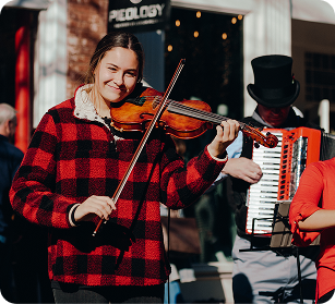 A young woman in a red and black plaid shirt plays the violin on the street, accompanied by a man in a top hat playing the accordion in Williamson County, Tennessee.