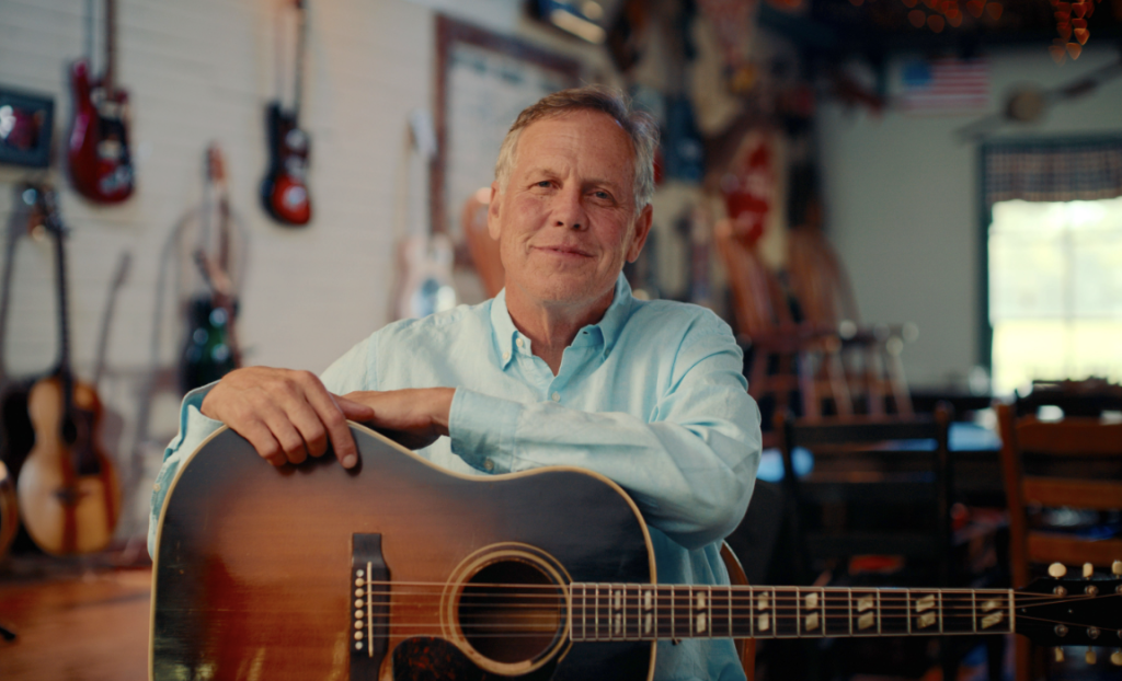 A man in a light blue shirt holds an acoustic guitar in a room filled with various musical instruments in Williamson County, Tennessee.