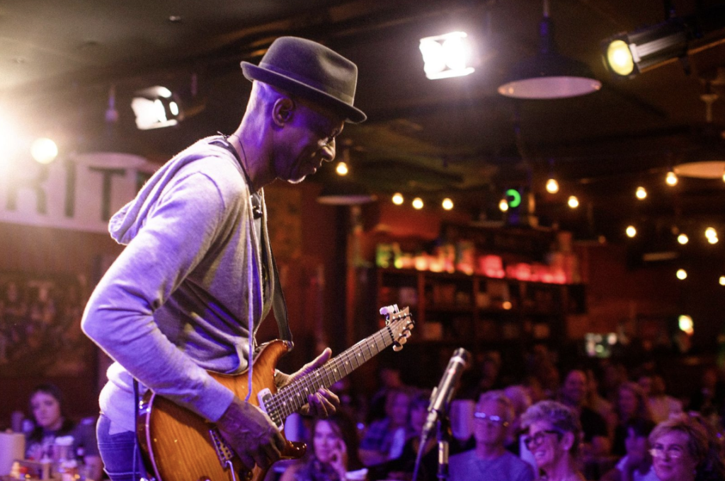 A musician wearing a hat plays an electric guitar on stage in a dimly lit venue in Williamson County, Tennessee.