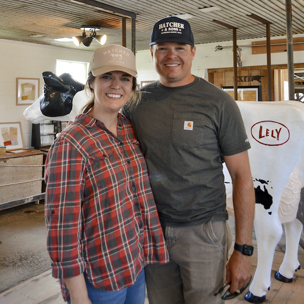 A smiling couple stands in a barn with a cow statue, wearing casual farm attire in Williamson County, Tennessee.
