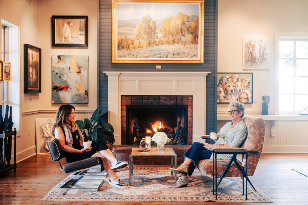 Two women enjoy tea by a cozy fireplace in an art-filled living room in Williamson County, Tennessee.