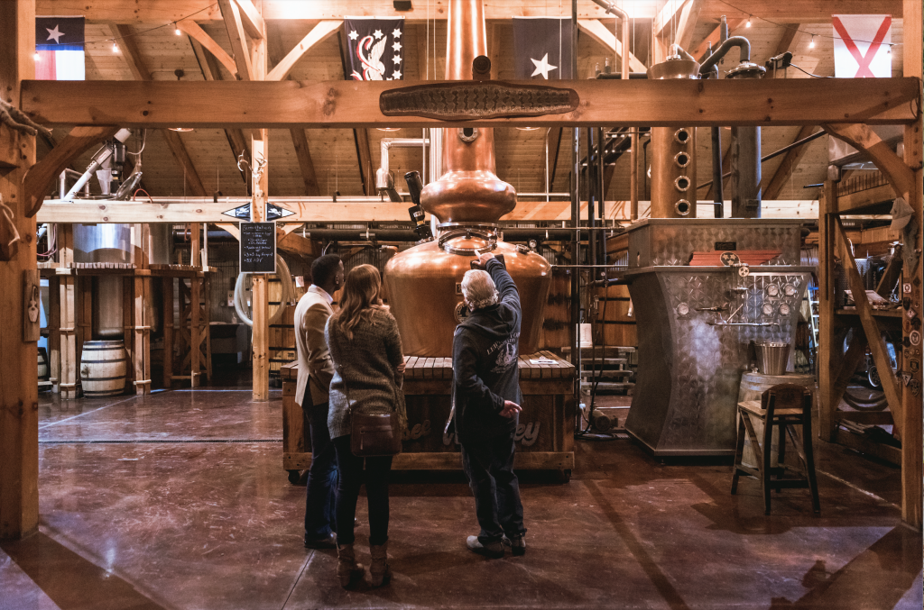 Three people tour a distillery, examining a large copper still in a wooden-beamed warehouse in Williamson County, Tennessee.