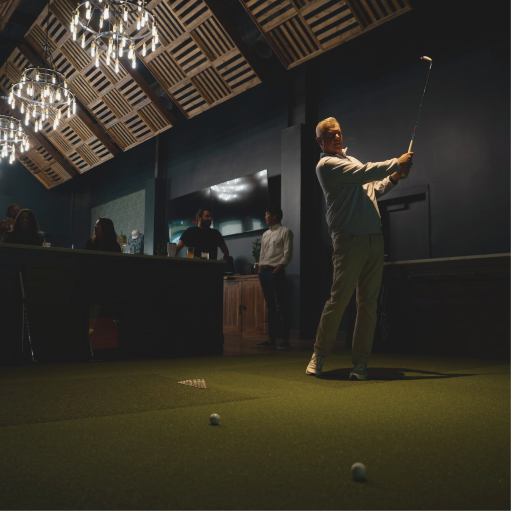 Man in golf attire swings club indoors, surrounded by onlookers and chandeliers in Williamson County, Tennessee.